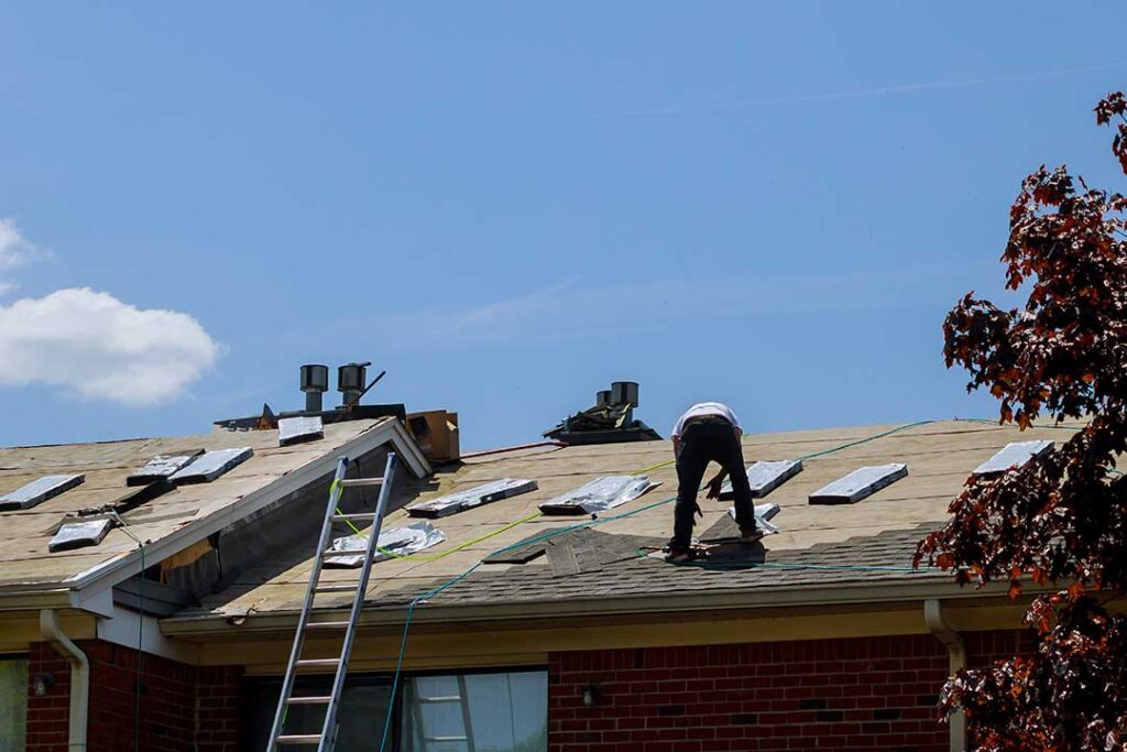 Homeowner examines roof shingles and gutters from a ladder beside a residential house.