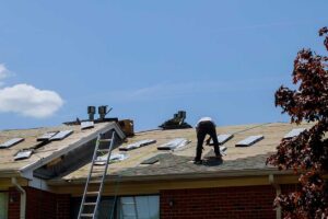 Homeowner examines roof shingles and gutters from a ladder beside a residential house.